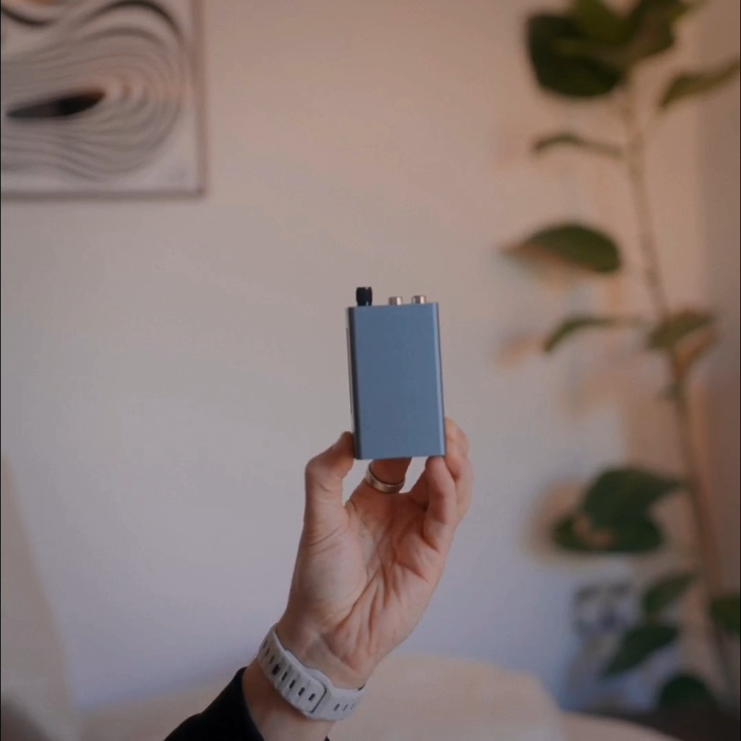 A hand holding a compact aluminium audio device with RCA outputs, showing how small it is